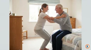 Caregiver aiuta anziano ad alzarsi dal letto con postura corretta e sicura.