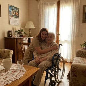 Una giovane caregiver abbraccia con affetto un’anziana signora su una sedia a rotelle
