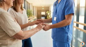 Infermiere in divisa blu stringe le mani di una donna anziana, supportata a sua volta da una donna più giovane, in un corridoio d'ospedale luminoso e moderno.