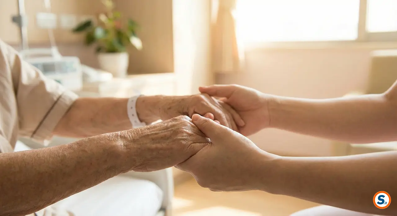 Primo piano delle mani di una persona anziana con braccialetto ospedaliero, tenute dolcemente dalle mani di un familiare o caregiver in una stanza luminosa.